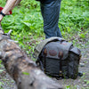 Nessmuk Pack sits on the ground next to a person sawing a tree. 