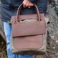 Person holding a brown leather and waxed canvas bag with a visible brand logo agaisnt a stone background. 