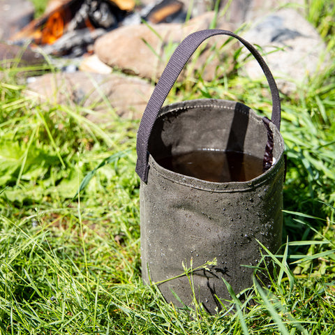 Waxed canvas bucket filled with water sits in the grass next to a fire pit. 